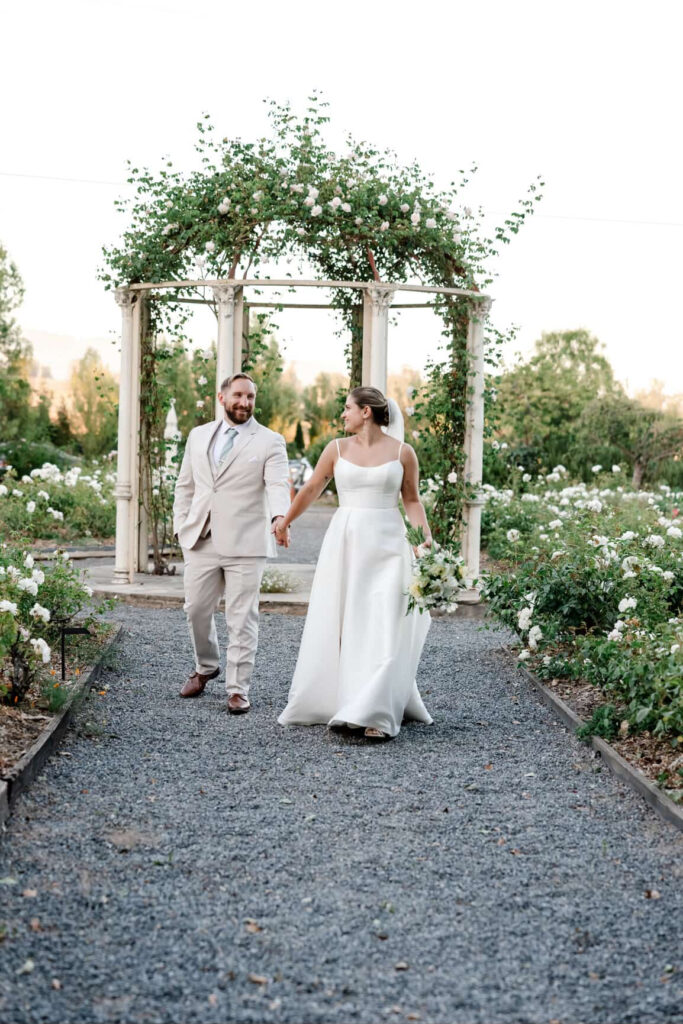 Bride and groom walking through the rose garden at Garden Valley Ranch during golden hour, holding hands and smiling.