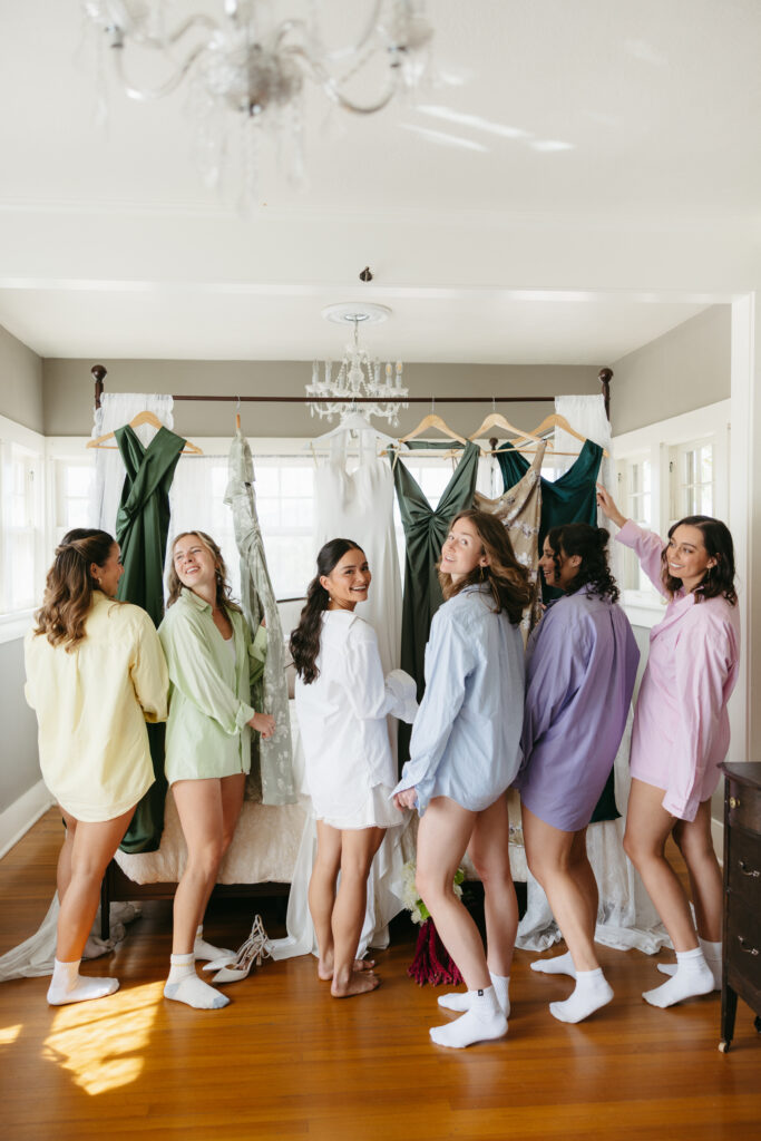 Bride and bridesmaids laughing in getting-ready room with emerald bridesmaid dresses hanging behind them