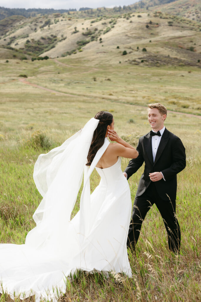 First look moment in Colorado foothills meadow — bride in strapless ballgown with cathedral veil walking toward groom in black tuxedo