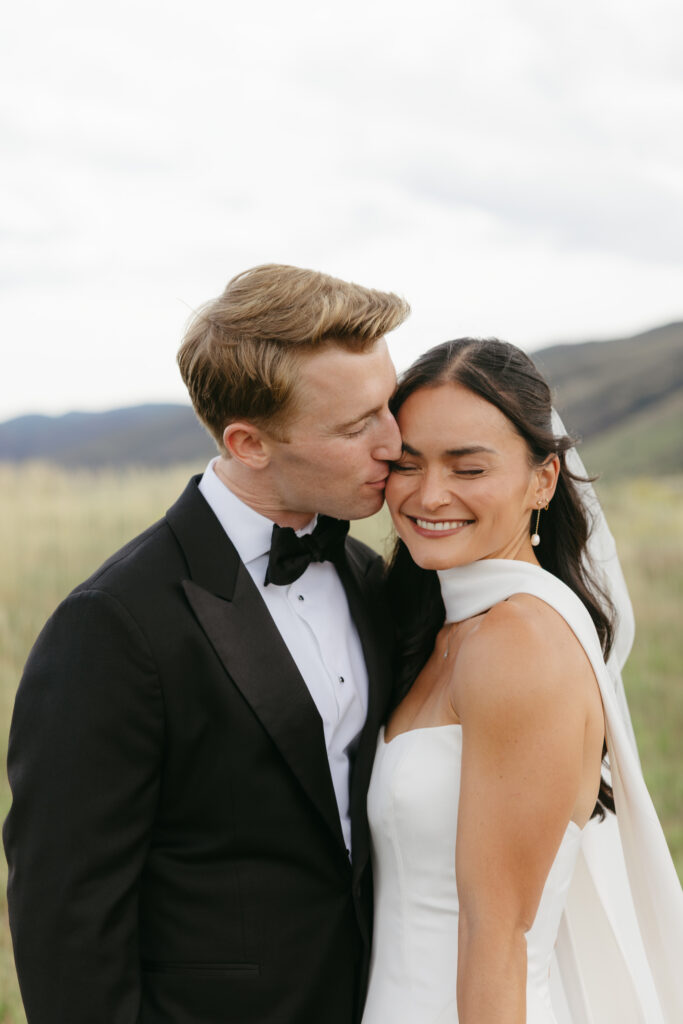 Groom in black tuxedo kissing bride on cheek during Colorado foothills first look portrait