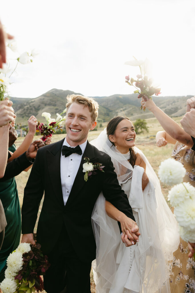 Colorado mountain wedding recessional — couple smiling as wedding party cheers around them with bouquets raised