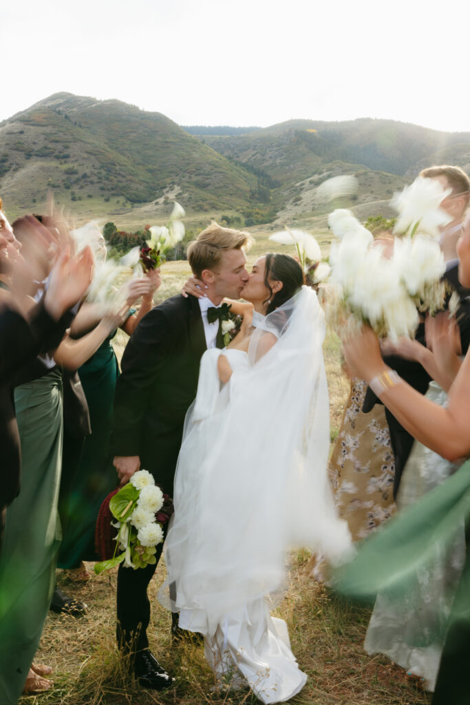 Bride and groom kissing surrounded by cheering wedding party on Colorado foothills hillside after ceremony