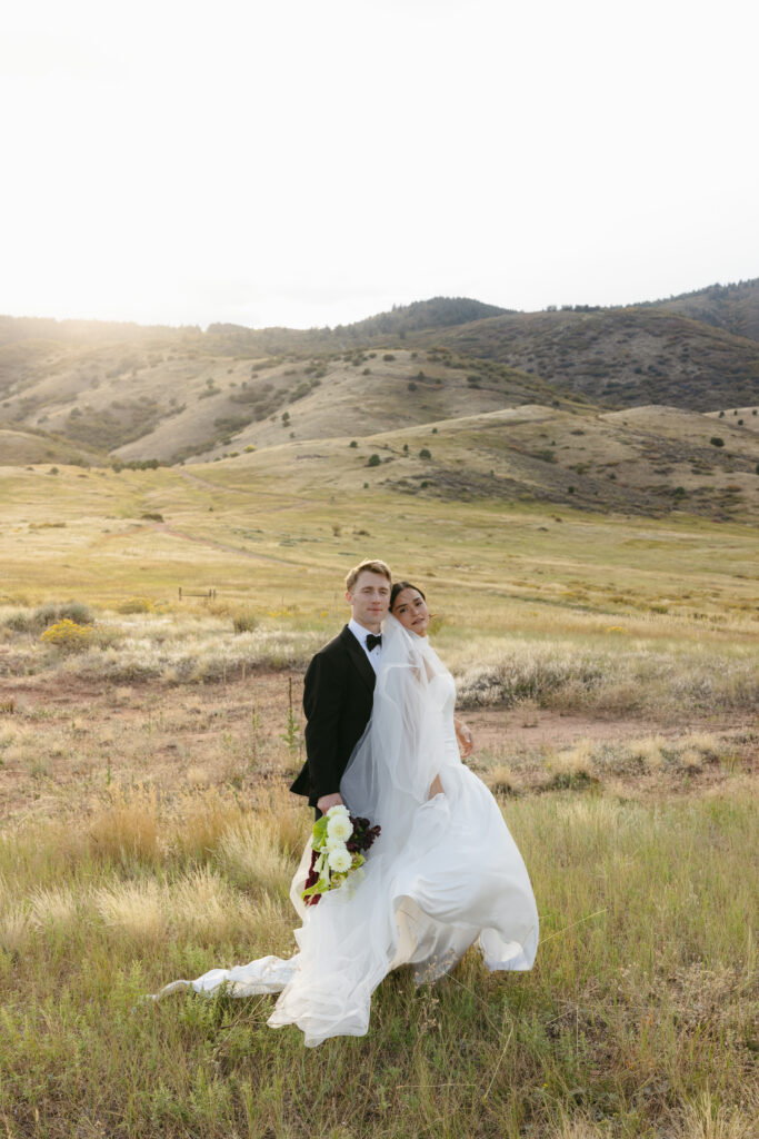 Bride and groom back-to-back in golden-hour Colorado foothills meadow with veil and train flowing in the wind
