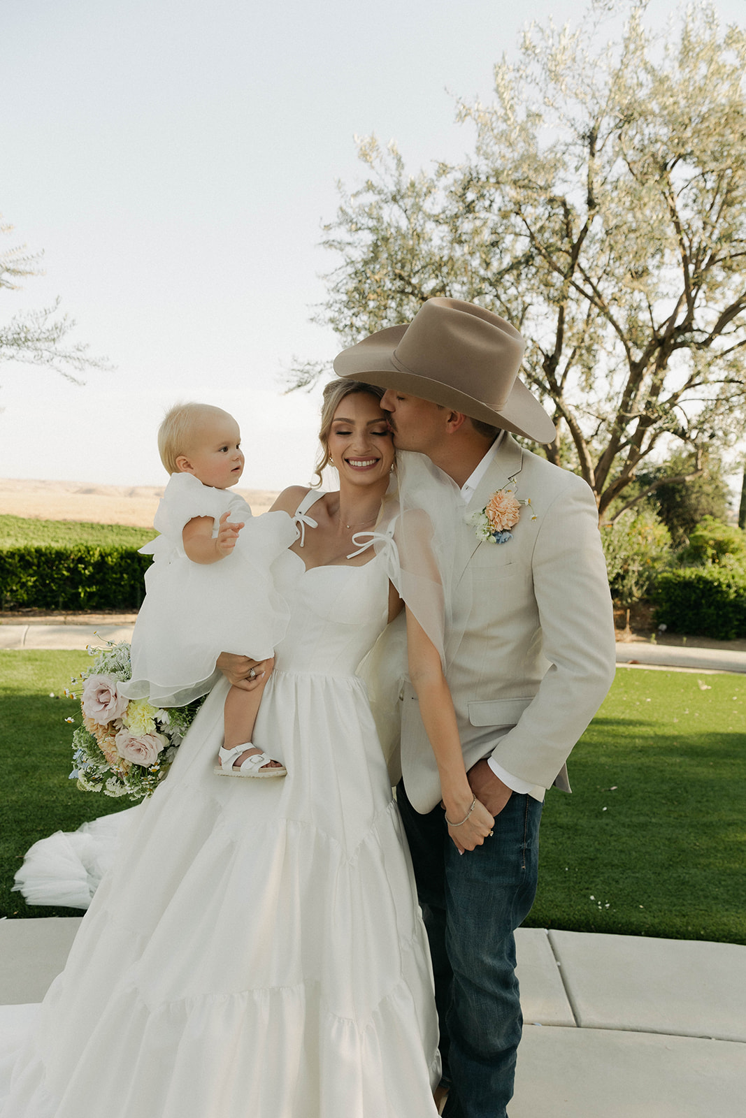 Groom in a cowboy hat kissing the bride while she holds their daughter