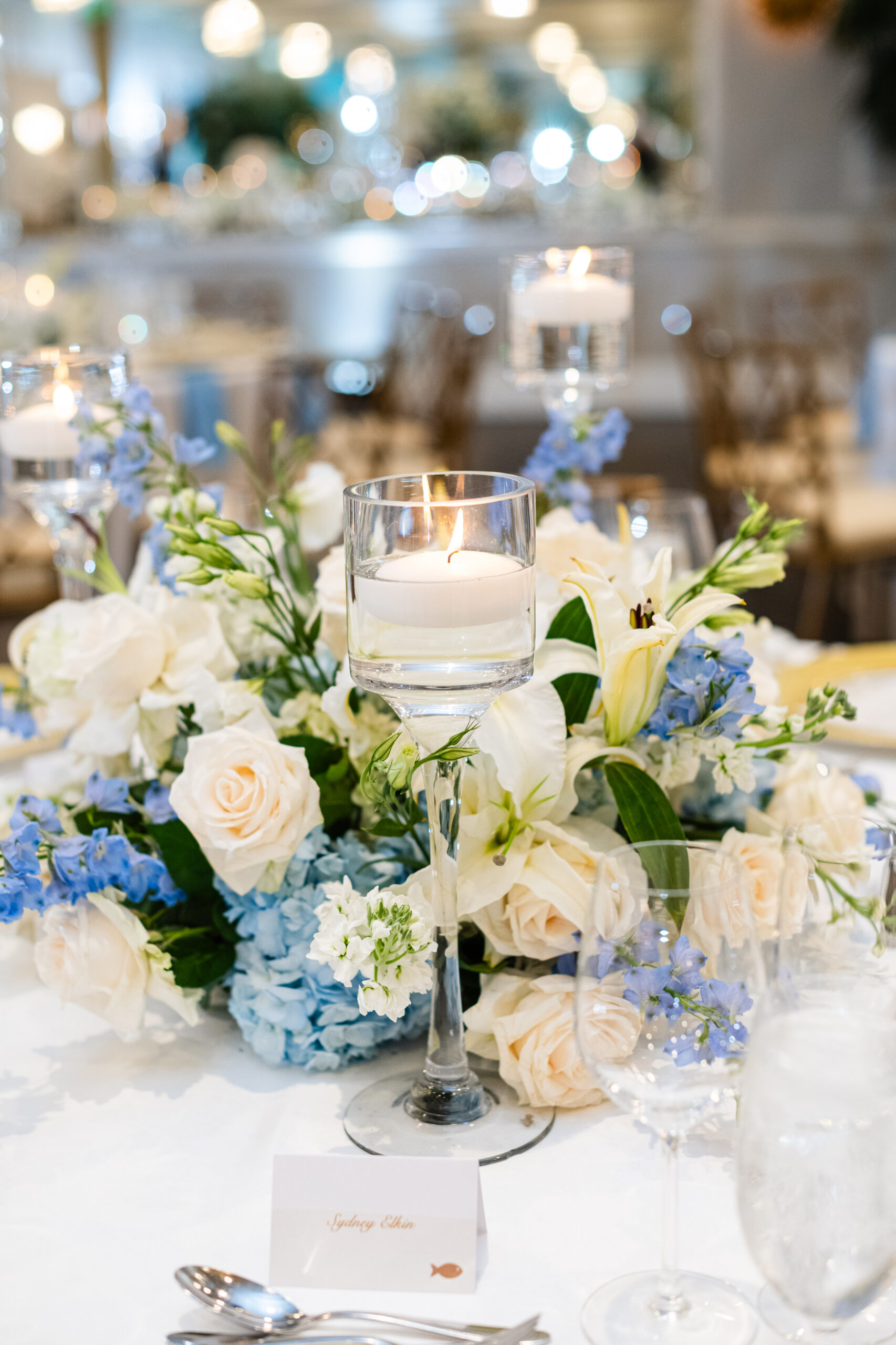 Centerpiece of white roses, lilies, and soft blue delphinium with a floating candle