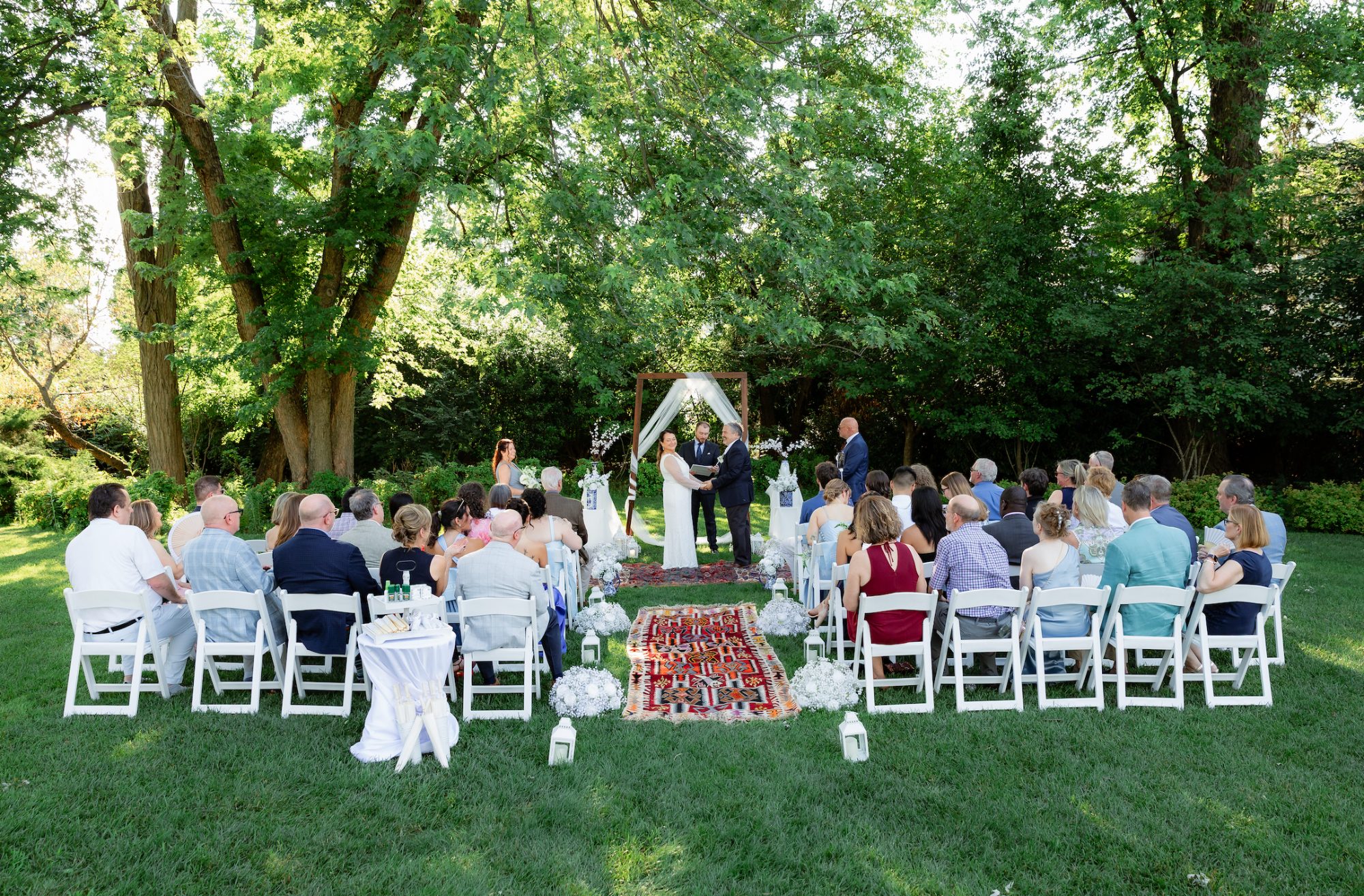 Garden wedding ceremony under tall trees at Rock Creek Mansion in Bethesda, Maryland with a Turkish rug aisle