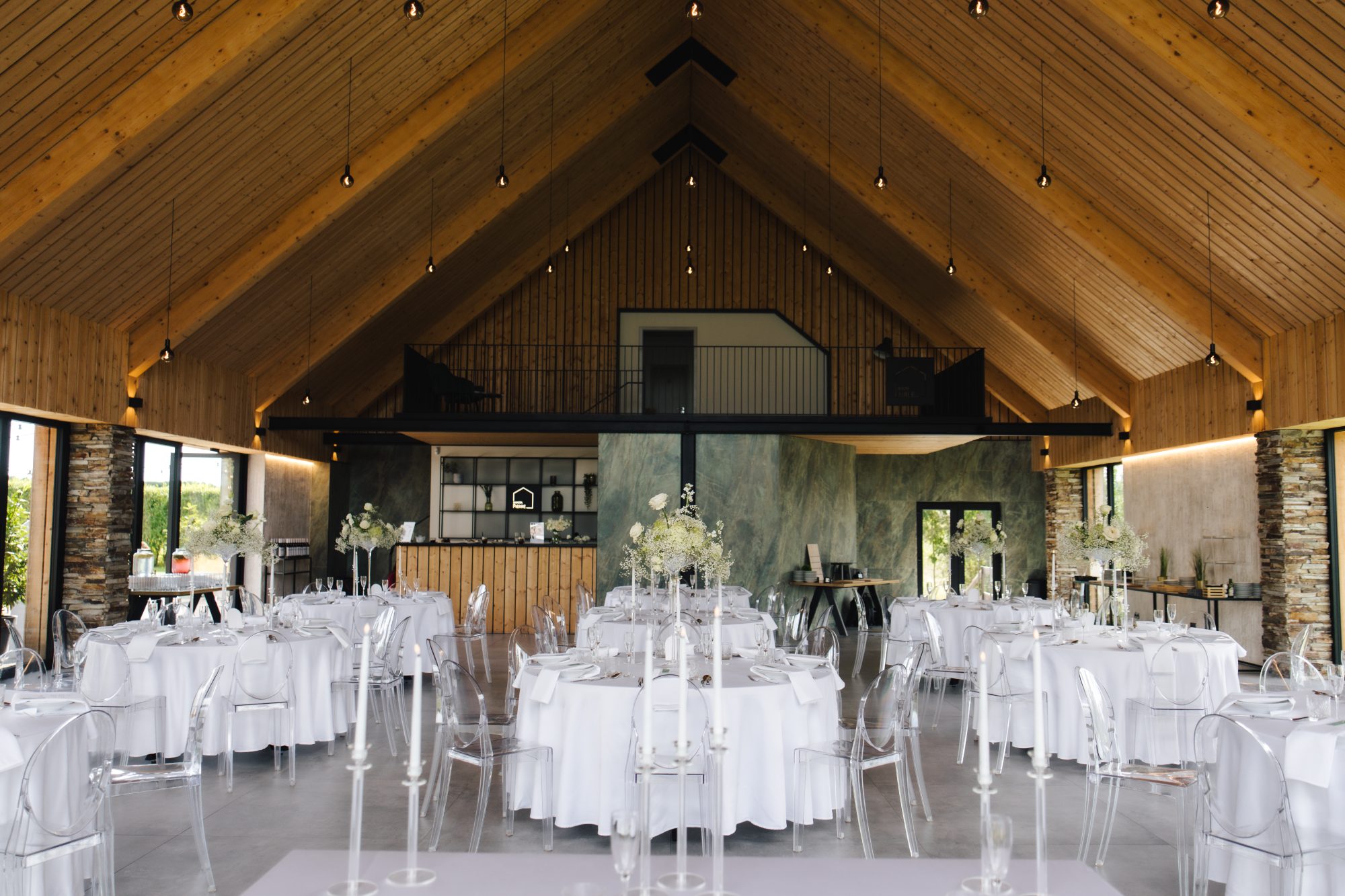 Round white tables and clear chairs inside the modern timber wedding hall